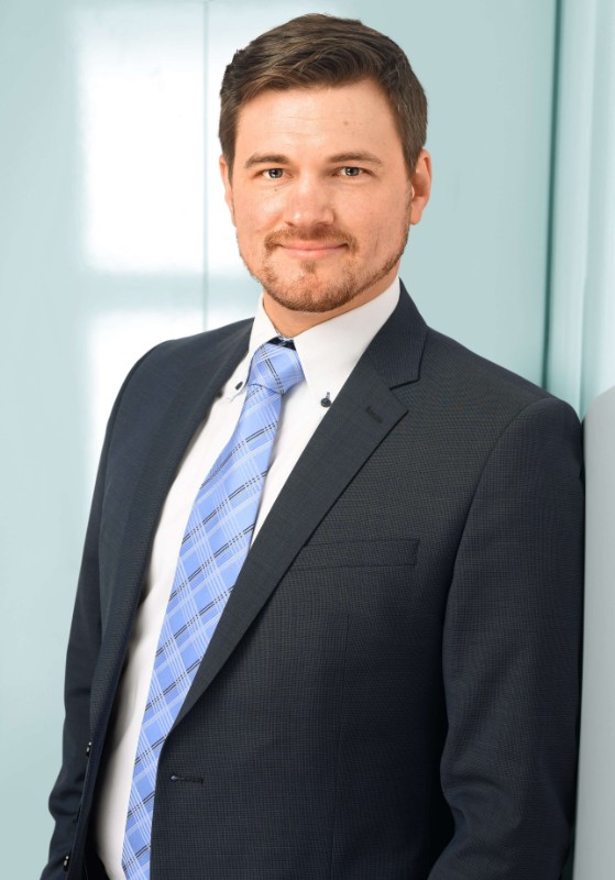 Portrait eines netten jungen Mannes mit kurzen braunen Haaren und Anzug, der vor hellblauem Hintergrund lächelt.
